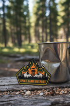 Metal camping pot with Spike Camp Outpost logo on a wooden surface in a forest setting